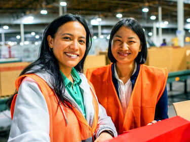 Dos mujeres sonriendo en un almacén con chalecos naranjas